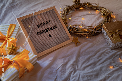 Felt letter board "Merry Christmas" on the bed decorated with golden wreath, lights and gift boxes