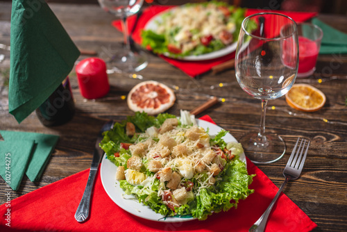 Christmas table with appetizing green Caesar salad with lettuce leaves and a glass on a red napkin on a wooden table