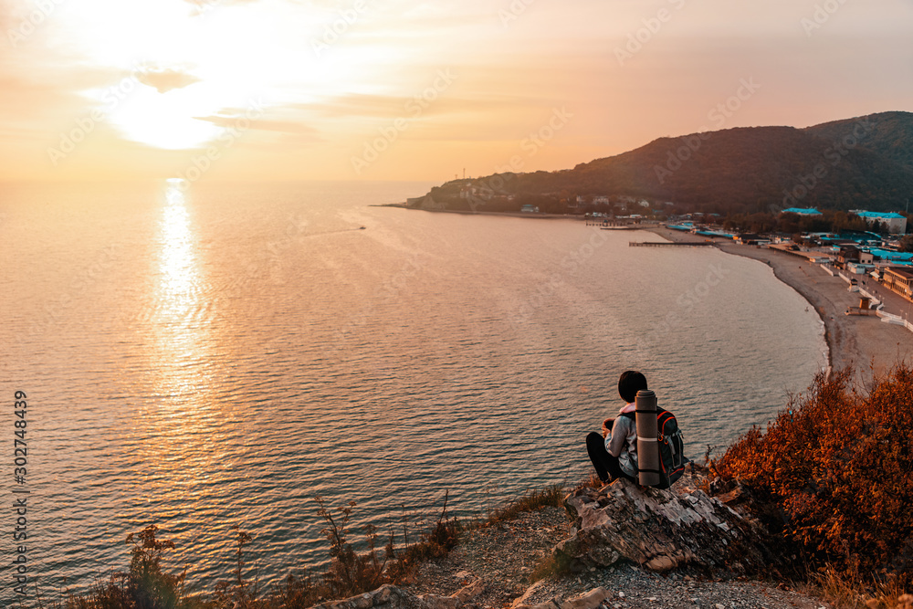 Tourism and sports recreation. A young woman with a tourist backpack on her back sits on a rock and looks at the sunset and the sea. View of the Bay. Copy space