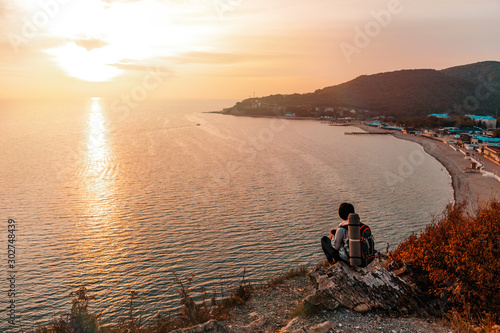 Tourism and sports recreation. A young woman with a tourist backpack on her back sits on a rock and looks at the sunset and the sea. View of the Bay. Copy space
