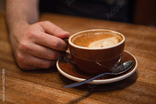 Male fingers holding brown cup of cappuccino on wooden table, Blurred background.