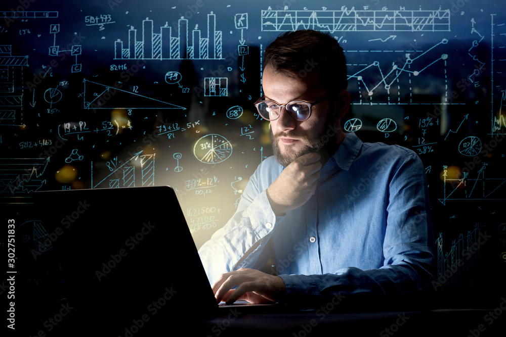 Young handsome businessman working late at night in the office with blue calculations in the background Young handsome businessman working late at night in the office with blue calculations in the background