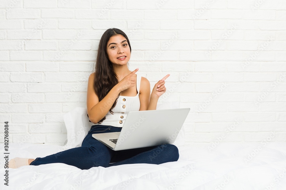 Young arab woman working with her laptop on the bed shocked pointing with index fingers to a copy space. Young arab woman working with her laptop on the bed shocked pointing with index fingers to a copy space.