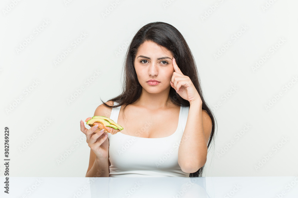 Young hispanic woman holding an avocado toast pointing his temple with finger, thinking, focused on a task. Young hispanic woman holding an avocado toast pointing his temple with finger, thinking, focused on a task.