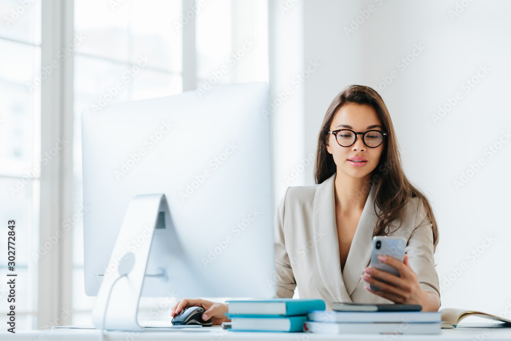 Elegant woman office worker uses mobile phone and computer at one time, sends messages and chats with clients online, monitors news from networks, sits at desktop with pile of notepads and books Elegant woman office worker uses mobile phone and computer at one time, sends messages and chats with clients online, monitors news from networks, sits at desktop with pile of notepads and books