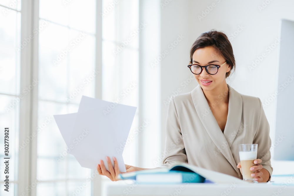 Horizontal view of confident brunette female worker in formal wear and optical spectacles looks through documentation, drink fresh beverage, focused down, poses indoor, has pleased expression Horizontal view of confident brunette female worker in formal wear and optical spectacles looks through documentation, drink fresh beverage, focused down, poses indoor, has pleased expression