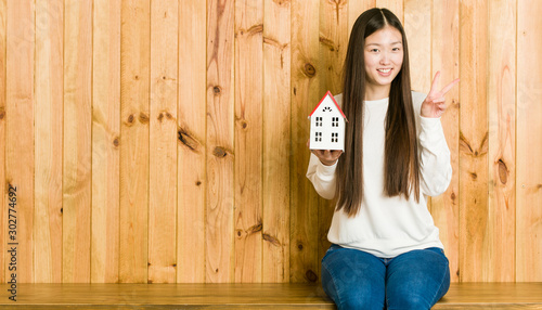 Young asian woman holding a house icon showing victory sign and smiling broadly. © Asier