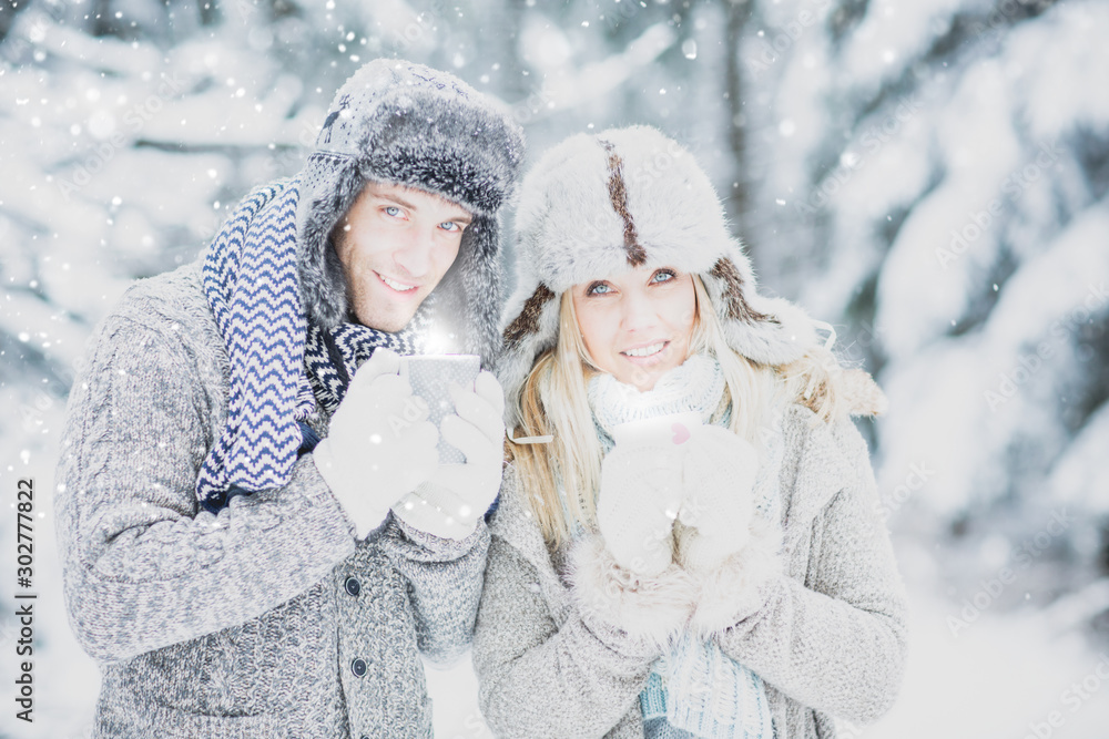 Young, pretty, couple in love with winter cap and scarf in winter landscape with cups and hot drink, warming up in the cold