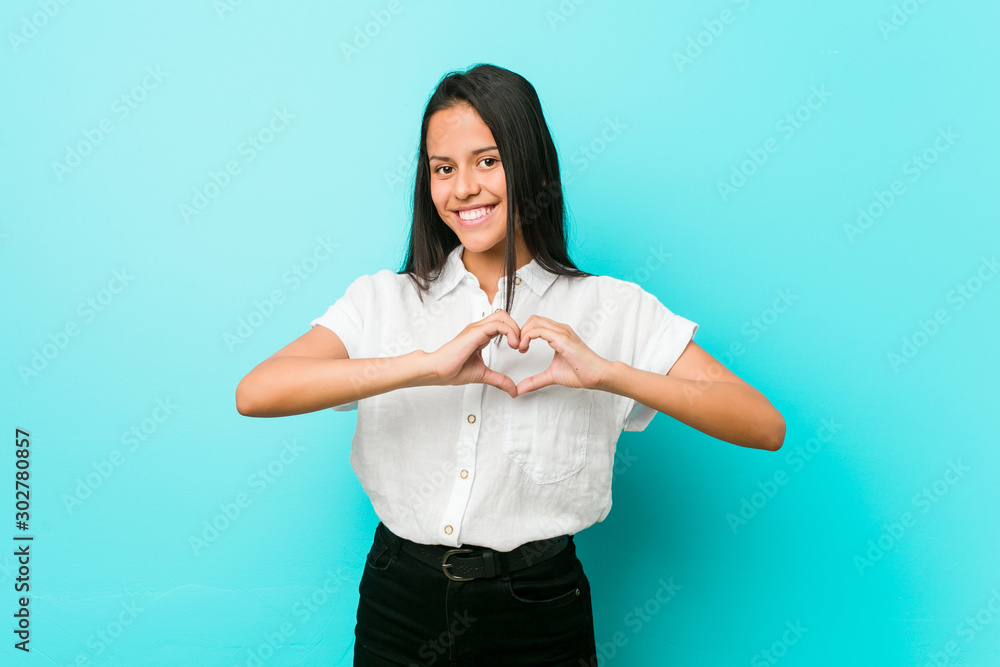 Young hispanic cool woman against a blue wall smiling and showing a heart shape with hands. Young hispanic cool woman against a blue wall smiling and showing a heart shape with hands.