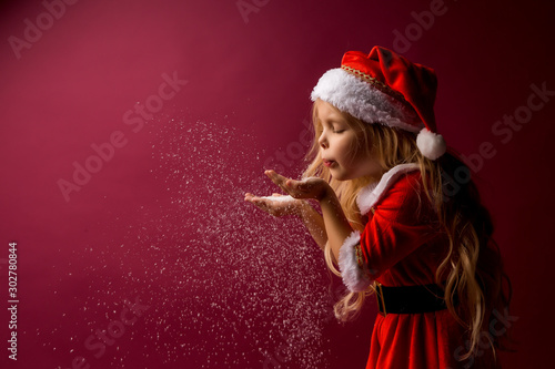 little blonde girl in a Santa suit blows snow off her hands. red background isolate. space for text