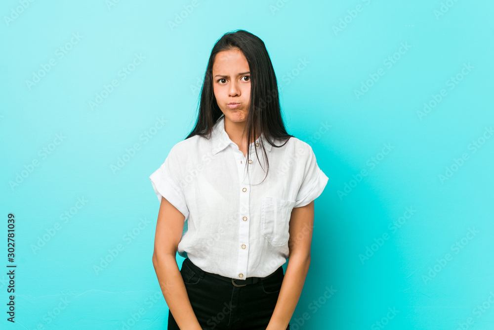Young hispanic cool woman against a blue wall blows cheeks, has tired expression. Facial expression concept. Young hispanic cool woman against a blue wall blows cheeks, has tired expression. Facial expression concept.