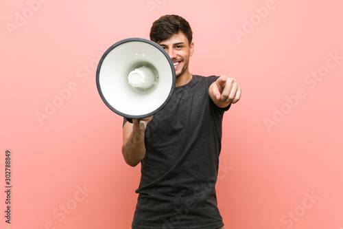 Young hispanic man holding a megaphone cheerful smiles pointing to front.