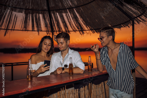 Smiling couple having fun using a smartphone in the beach bar, while their friend has a call on a mobile phone