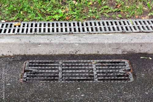 sewer manhole grille on an asphalt road near the curb in the background behind the curb a drain channel covered with a grill.
