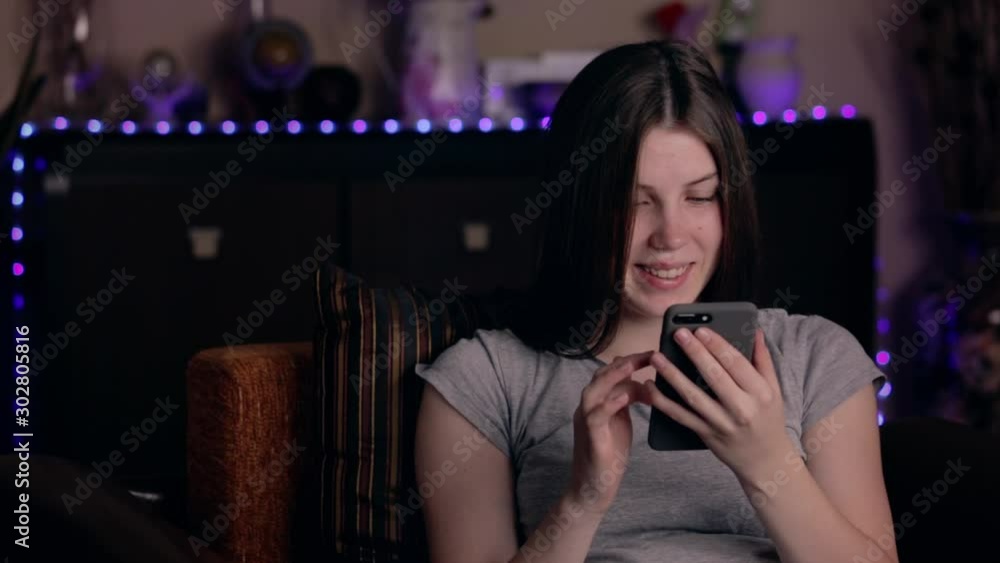 Beautiful young woman sitting on the couch at home looking at phone with a smile and typing a message Beautiful young woman sitting on the couch at home looking at phone with a smile and typing a message