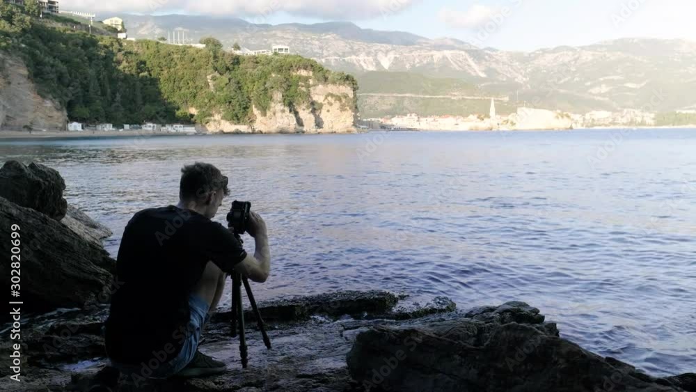 A man setting up his camera to photograph Budva Old town in Montenegro with waves crashing in front of him during sunset.