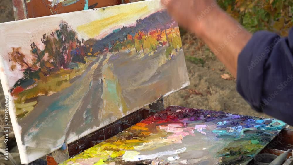 Oil painting on canvas. Professional artist adult man paints a picture in nature. Painting in the open air using a portable easel and a paintbox Oil painting on canvas. Professional artist adult man paints a picture in nature. Painting in the open air using a portable easel and a paintbox
