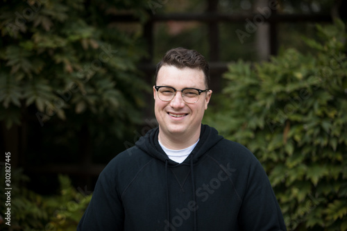 A caucasian male wearing a black hoodie sweater and glasses poses in a garden while surrounded by fall foliage on a cold afternoon.