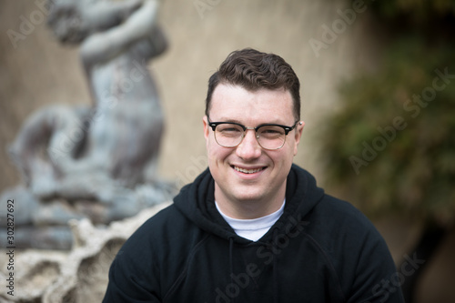 A caucasian male wearing a black hoodie sweater and glasses poses in a garden while surrounded by fall foliage on a cold afternoon.