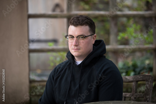 A caucasian male wearing a black hoodie sweater and glasses poses in a garden while surrounded by fall foliage on a cold afternoon.
