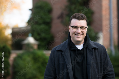 A caucasian male wearing a black hoodie sweater and glasses poses in a garden while surrounded by fall foliage on a cold afternoon.