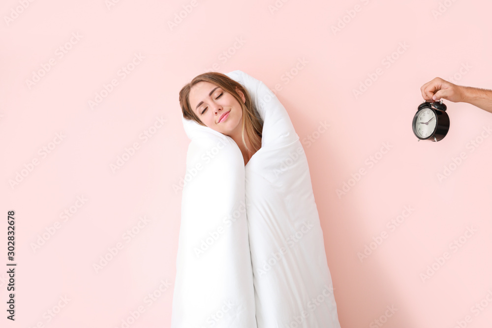 Sleepy young woman wrapped in blanket and male hand with alarm clock on color background - obrazy, fototapety, plakaty 