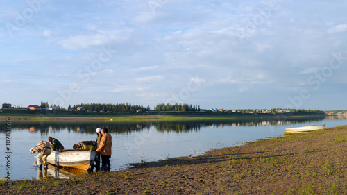 A group of Yakuts sits in a boat standing at the Bank of the river Vilyuy before sailing to the shore with the houses of the village at sunset.