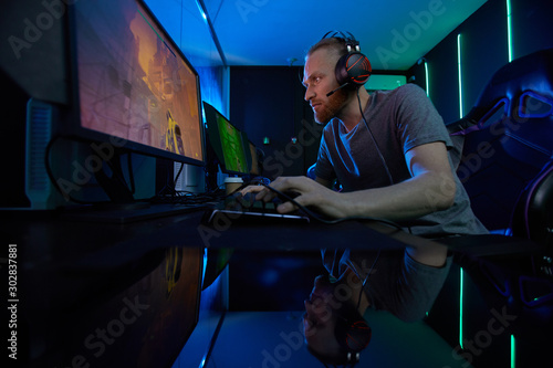 Young bearded computer gamer in headphones concentrating on his game he sitting in gaming chair and looking at computer monitor in dark room