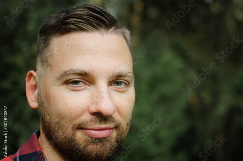 Close up portrait of young bearded man in red shirt looking at the camera with smile in the park on trees background