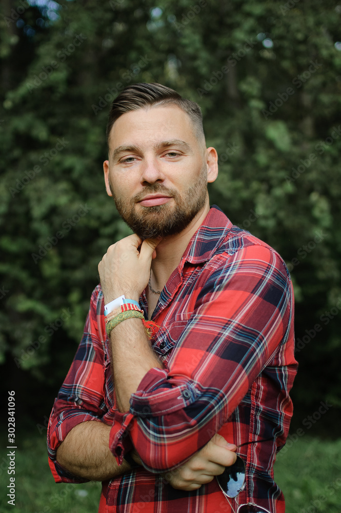 Brutal bearded young man in red shirt is standing outdoors on green trees background. Hipster style concept. Brutal bearded young man in red shirt is standing outdoors on green trees background. Hipster style concept.