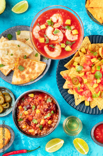 Mexican food, many dishes of the cuisine of Mexico, flat lay top-down shot from on a vibrant blue background. Shrimp cocktail, nachos, tequila, chili con carne, quesadillas