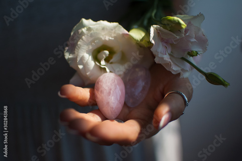 Female hands holding two yoni eggs for vumfit, imbuilding or meditation are made from pink quartz and transparent violet amethyst with white flowers indoors photo