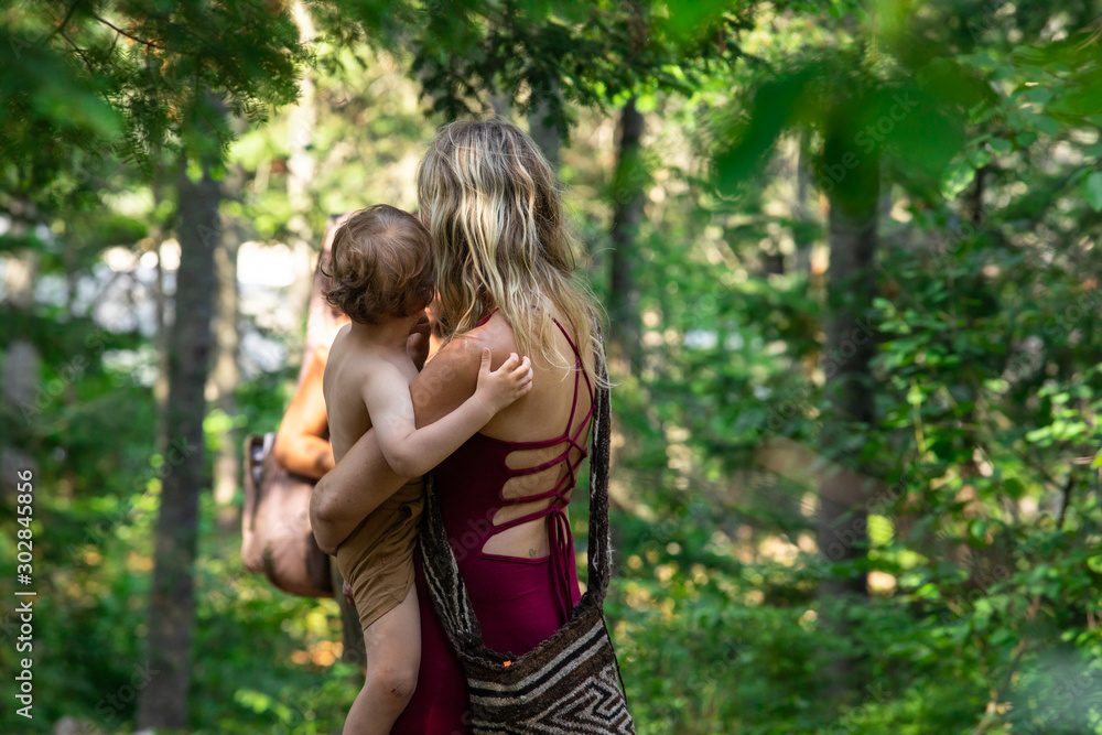 A young mother holds her child in her arms viewed from the back as they walking through a woodland trail with family and friends A young mother holds her child in her arms viewed from the back as they walking through a woodland trail with family and friends