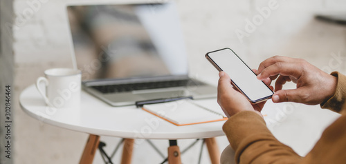 Close-up view of young man holding blank screen smartphone with laptop computer and office supplies