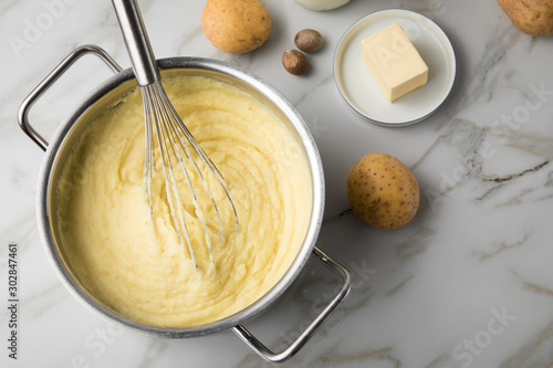 Kochtopf mit Kartoffelpüree mit Zutaten aus Kartoffeln, Milch, Salz, Butter, Muskatnuss dazu Muskatreibe und Schneebesen auf hell Marmor Hintergrund photo