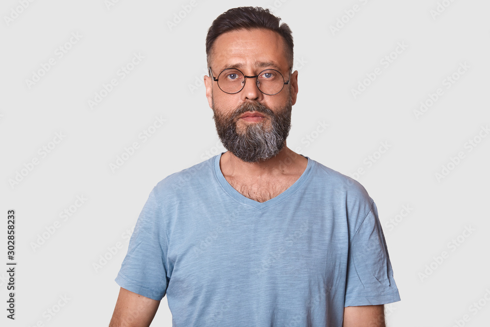 Close up portrait of middle aged male wearing eyeglasses and caual gray t shirt, looking directly at camera, posing against white background with copy space, attractive guy with beard. People concept. Close up portrait of middle aged male wearing eyeglasses and caual gray t shirt, looking directly at camera, posing against white background with copy space, attractive guy with beard. People concept.