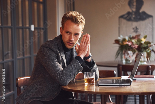 Elegant young guy in formal wear sits in cafe with his laptop and glass of alcohol photo