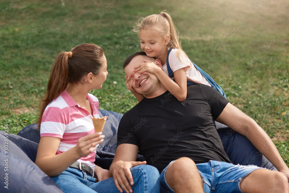 Family portrait of mommy, daddy, funny and cute daughter, female child covering dad's eyes with her palms, people wearing casual clothing, spending time together in open air. Happyness concept.