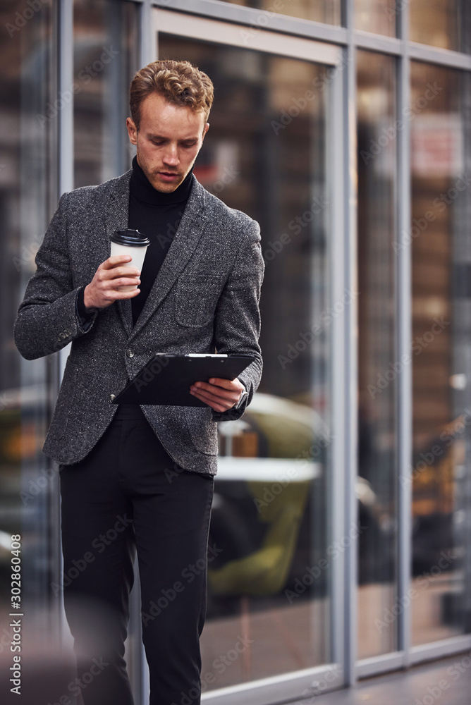 Man in elegant formal wear with cup of drink and notepad in hands is outside against modern building Man in elegant formal wear with cup of drink and notepad in hands is outside against modern building