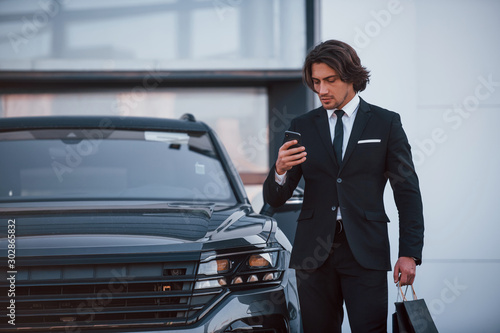 Portrait of handsome young businessman in black suit and tie outdoors near modern car and with shopping bags © standret