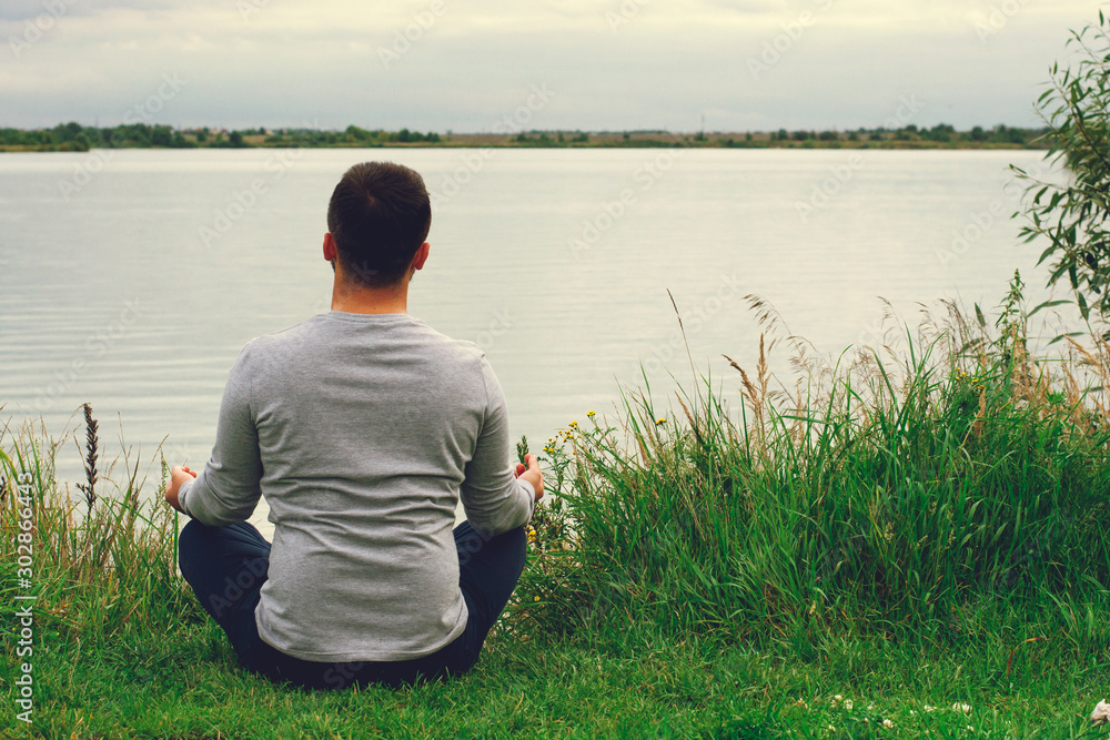 Fototapeta premium A young guy in a light polo sits on the shore. The view from the back. Yoga training. Appeasement.