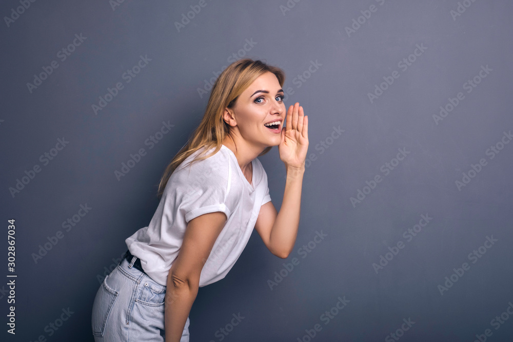 Caucasian woman in neutral casual outfit standing on a neutral grey background. Portrait with emotions: happiness, amazement, joy and satisfaction while whispering secrets Caucasian woman in neutral casual outfit standing on a neutral grey background. Portrait with emotions: happiness, amazement, joy and satisfaction while whispering secrets