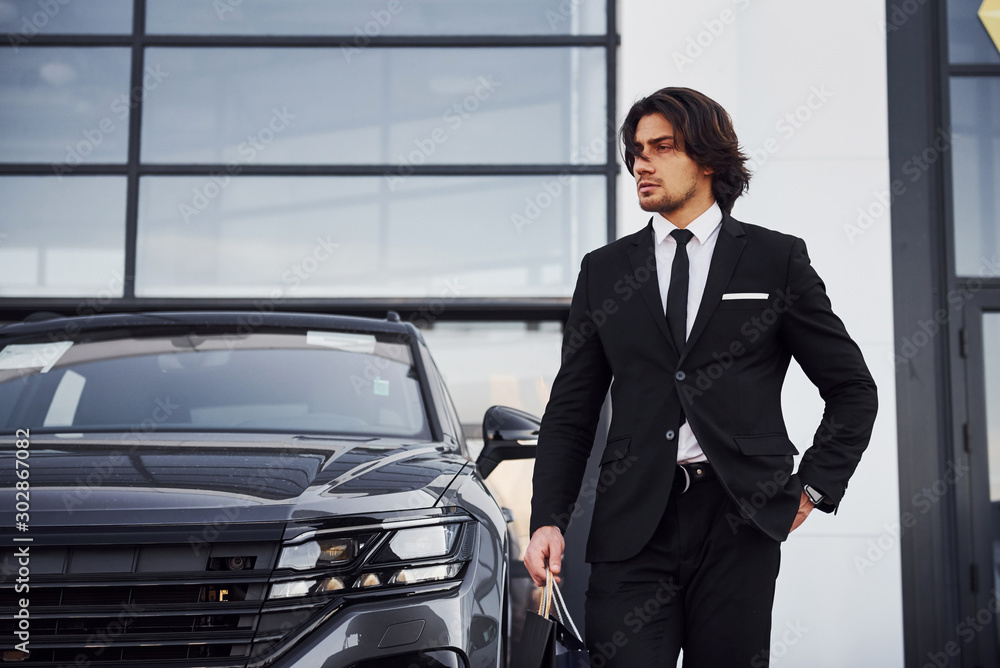 Portrait of handsome young businessman in black suit and tie outdoors near modern car and with shopping bags Portrait of handsome young businessman in black suit and tie outdoors near modern car and with shopping bags