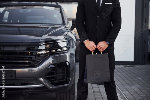 Close up view of businessman in black suit and tie stands near modern automobile with shopping bags in hands © standret