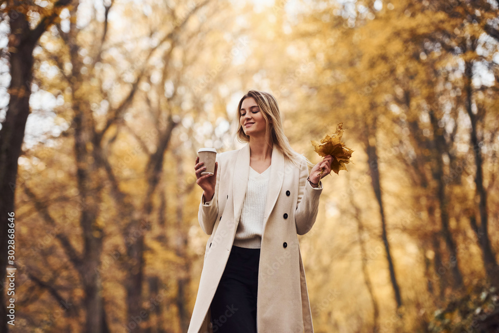 Holds cup of drink. Portrait of young brunette that is in autumn forest at daytime