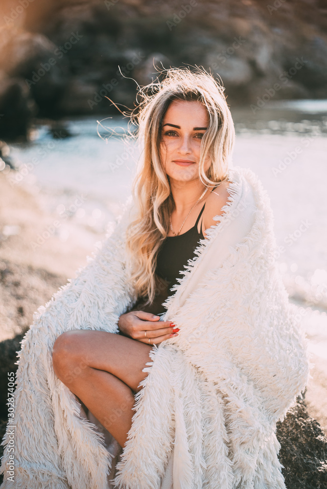 Beautiful girl with beautiful hair in shorts sitting on a rock on the background of rocks and the sea or ocean, wrapped in a fluffy light plaid Beautiful girl with beautiful hair in shorts sitting on a rock on the background of rocks and the sea or ocean, wrapped in a fluffy light plaid
