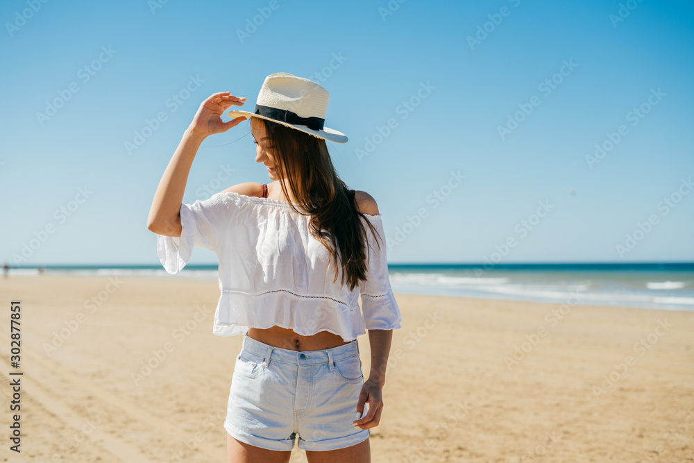 smiling girl looking away to the side and holding a hat on the beach with her hand