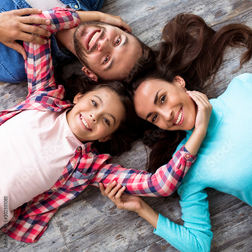 Lovely family. Close up photo of beautiful young family lying on the floor head to head, smiling broadly and hugging together.