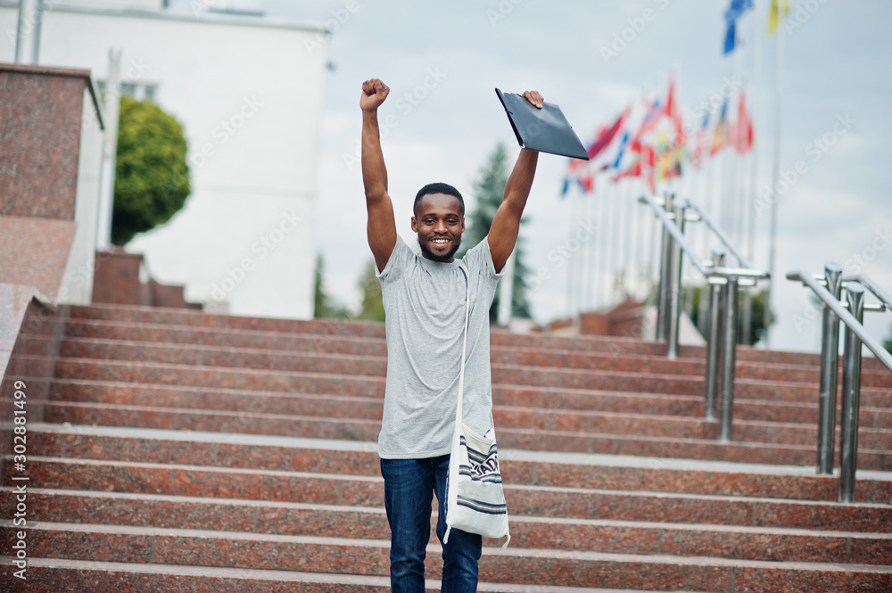 Hands up i am a winner! African student male posed with backpack and school items on yard of university, against flags of different countries.