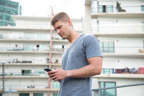 Focused young man using smartphone. Portrait of handsome concentrated young man standing on street and texting via cell phone. Technology concept